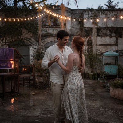an original couple dancing under string lights in a garden at dusk: a man with short black hair in a linen shirt and a woman with braided red hair in a summer dress