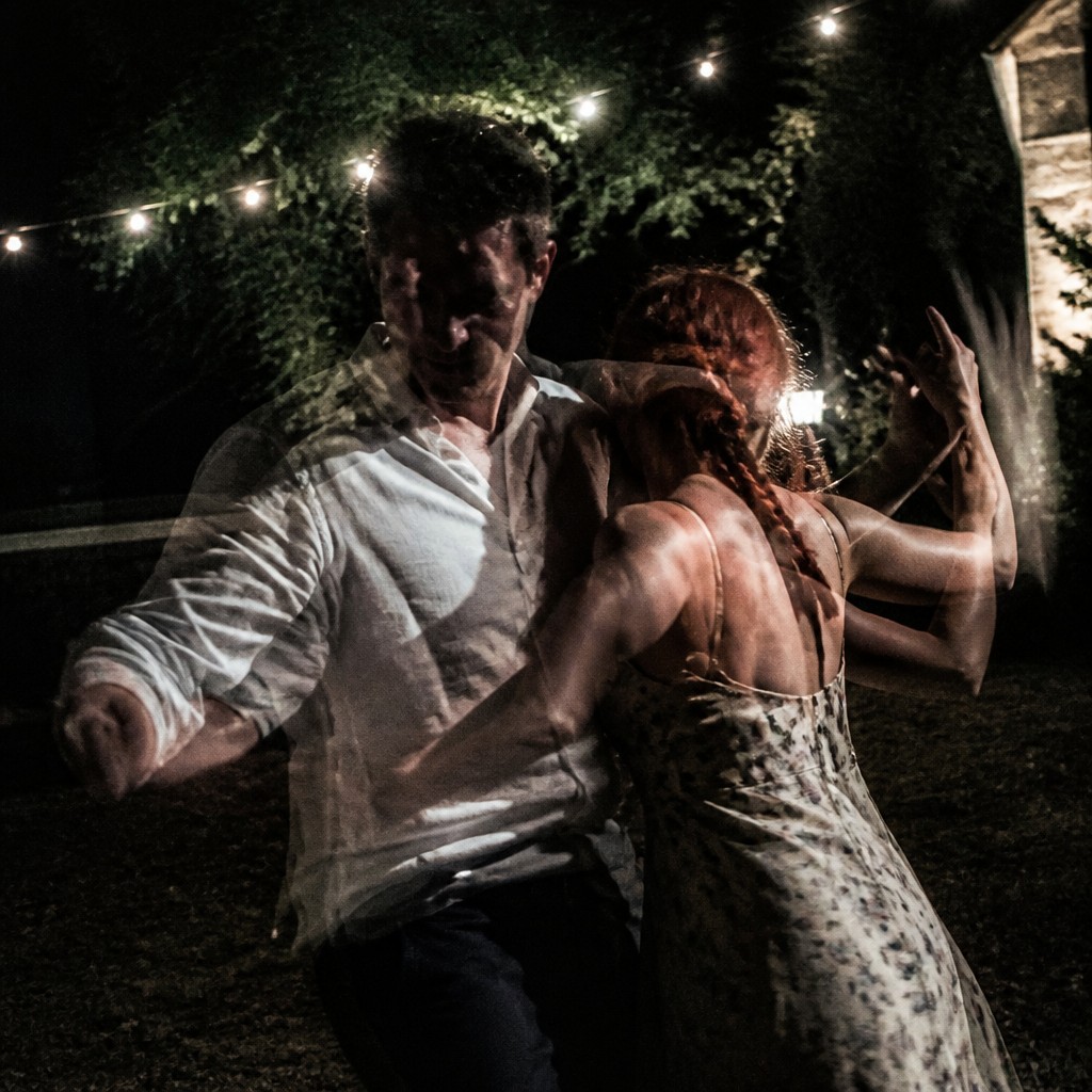 an original couple dancing under string lights in a garden at dusk: a man with short black hair in a linen shirt and a woman with braided red hair in a summer dress