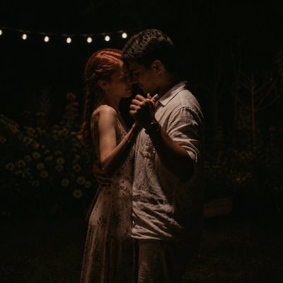 an original couple dancing under string lights in a garden at dusk: a man with short black hair in a linen shirt and a woman with braided red hair in a summer dress