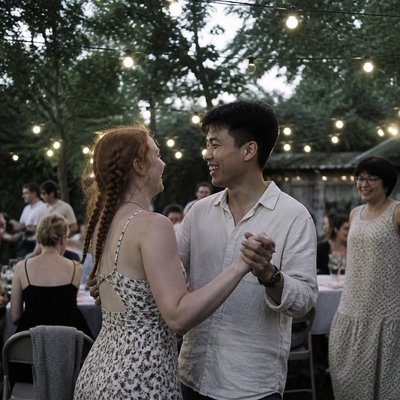 an original couple dancing under string lights in a garden at dusk: a man with short black hair in a linen shirt and a woman with braided red hair in a summer dress