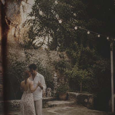 an original couple dancing under string lights in a garden at dusk: a man with short black hair in a linen shirt and a woman with braided red hair in a summer dress