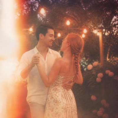 an original couple dancing under string lights in a garden at dusk: a man with short black hair in a linen shirt and a woman with braided red hair in a summer dress