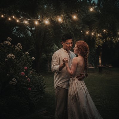 an original couple dancing under string lights in a garden at dusk: a man with short black hair in a linen shirt and a woman with braided red hair in a summer dress