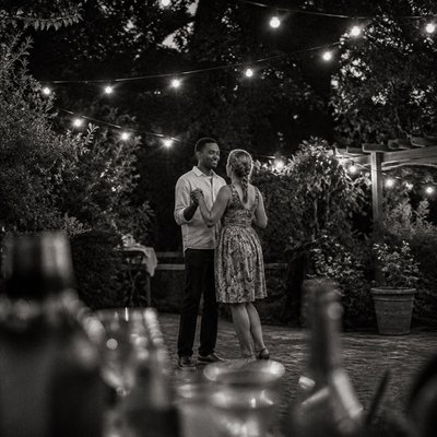 an original couple dancing under string lights in a garden at dusk: a man with short black hair in a linen shirt and a woman with braided red hair in a summer dress