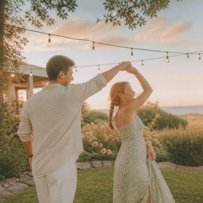 an original couple dancing under string lights in a garden at dusk: a man with short black hair in a linen shirt and a woman with braided red hair in a summer dress