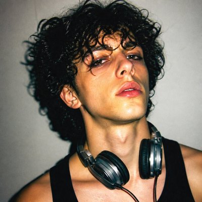 head-and-shoulders portrait of an original character: a young man with curly dark hair and headphones around his neck, plain backdrop, no text