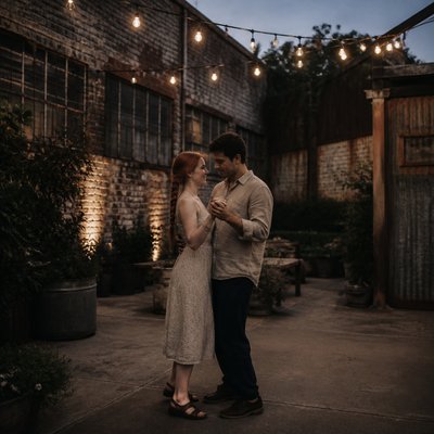 an original couple dancing under string lights in a garden at dusk: a man with short black hair in a linen shirt and a woman with braided red hair in a summer dress