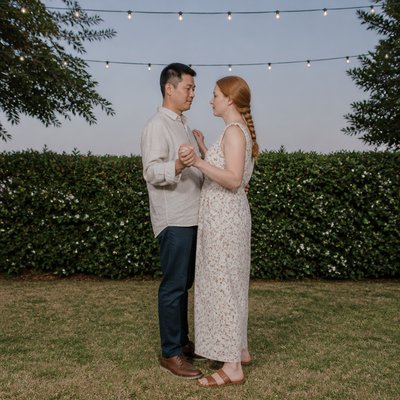 an original couple dancing under string lights in a garden at dusk: a man with short black hair in a linen shirt and a woman with braided red hair in a summer dress