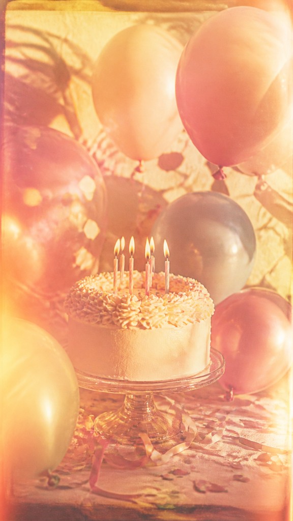 a birthday cake with lit candles surrounded by balloons and confetti, festive vertical composition, no text