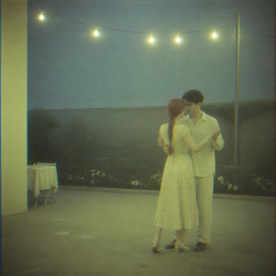 an original couple dancing under string lights in a garden at dusk: a man with short black hair in a linen shirt and a woman with braided red hair in a summer dress